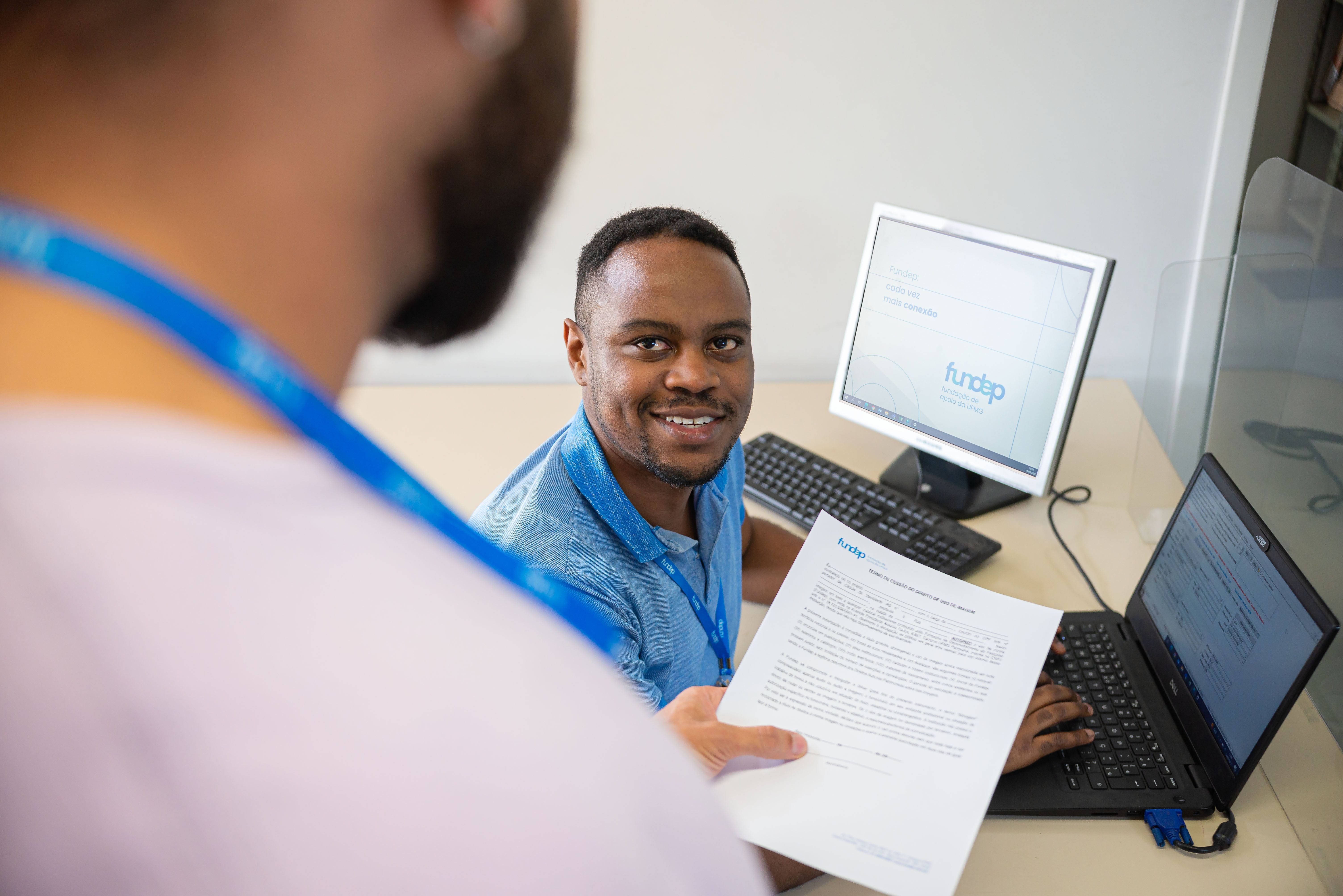 HOmem negro sorridente assentado em frente a duas telas de computador recebe um documento das mãos de um homem pardo que está em pé. 